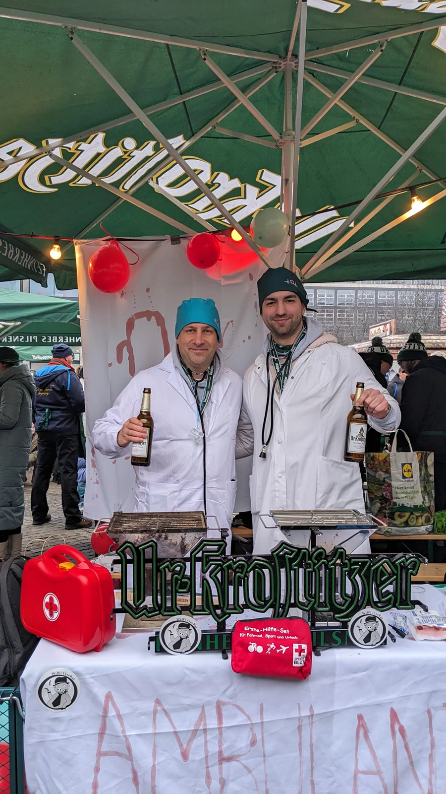 Zwei Männer in weißen Jacken stehen unter einem grünen Zelt und lächeln, einer hält eine Flasche.