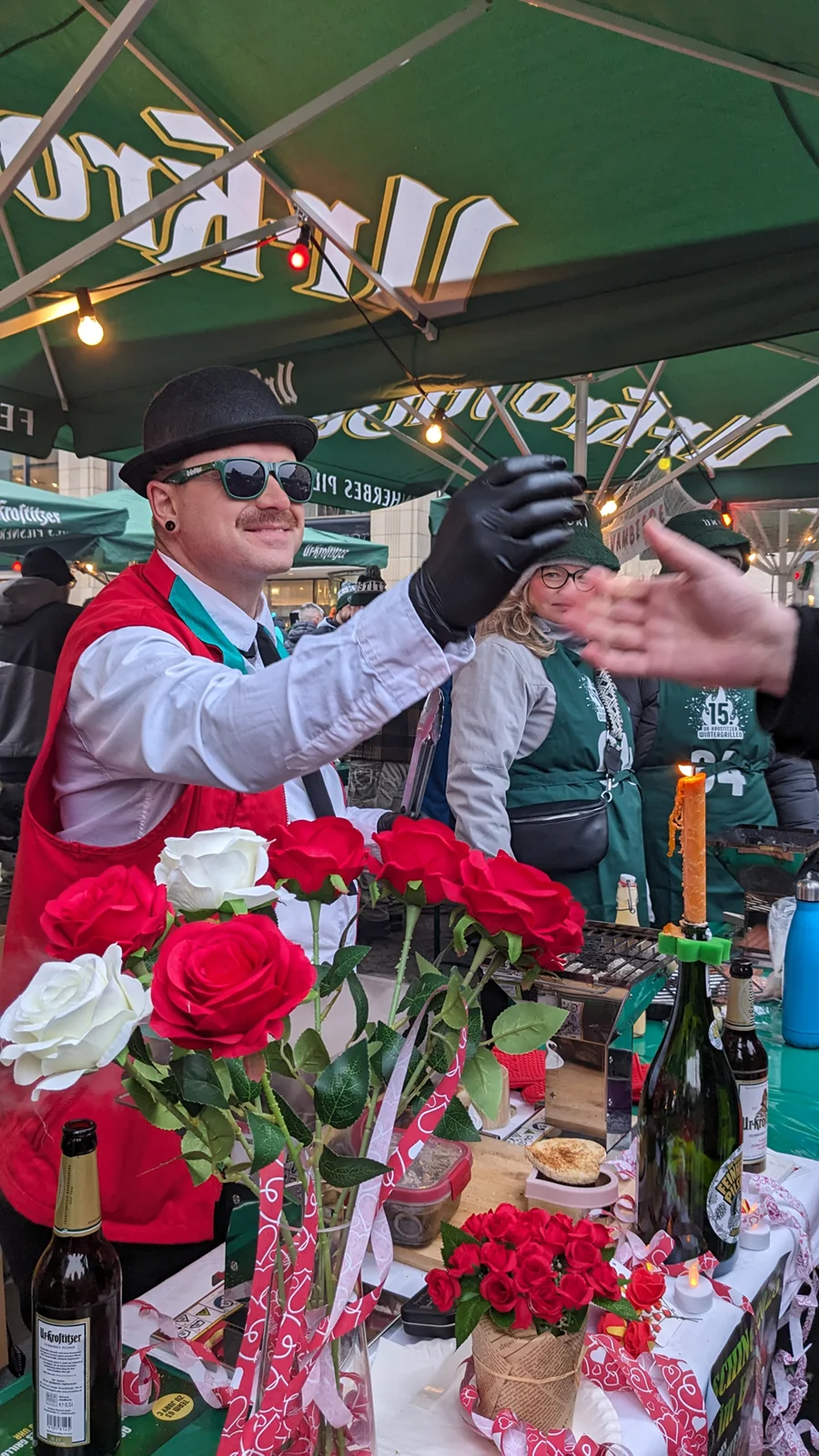 Ein Mann in Melone und roter Weste serviert Essen an einem dekorierten Marktstand.