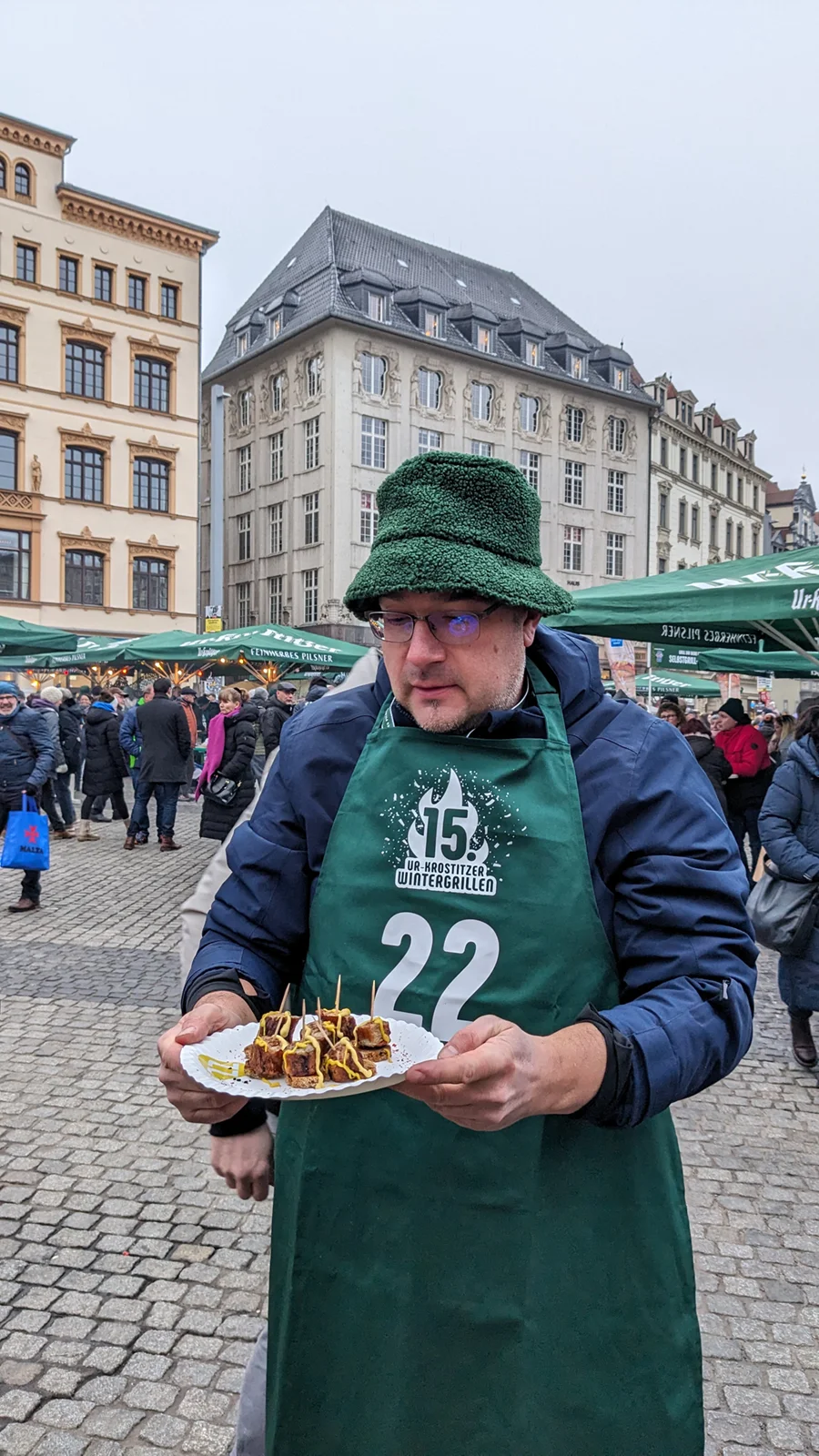 Ein Mann mit grüner Schürze hält einen Teller mit Snacks auf einem belebten Markt.