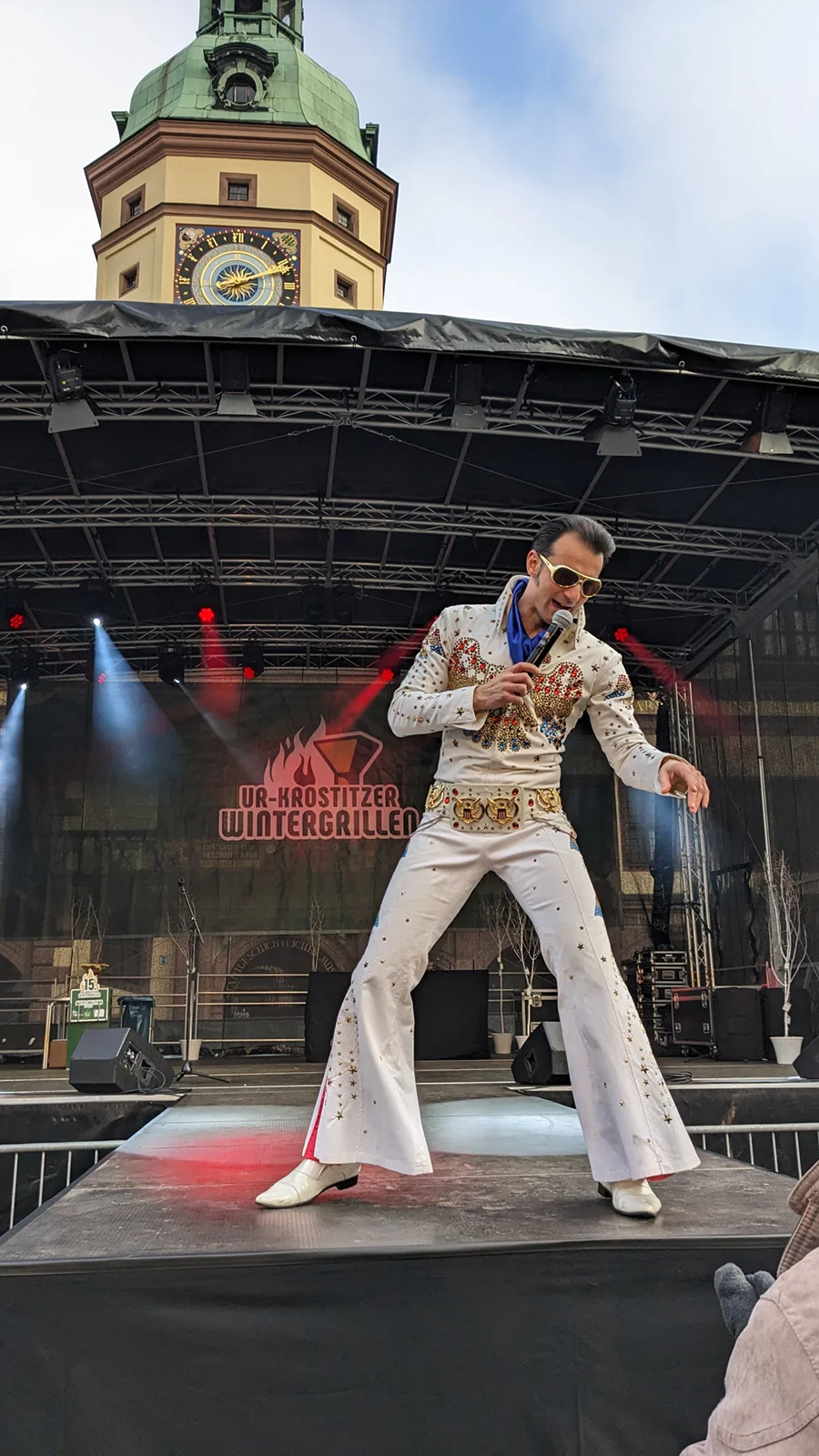 Ein Mann in weißem Glitzeranzug und Sonnenbrille singt auf einer Bühne.