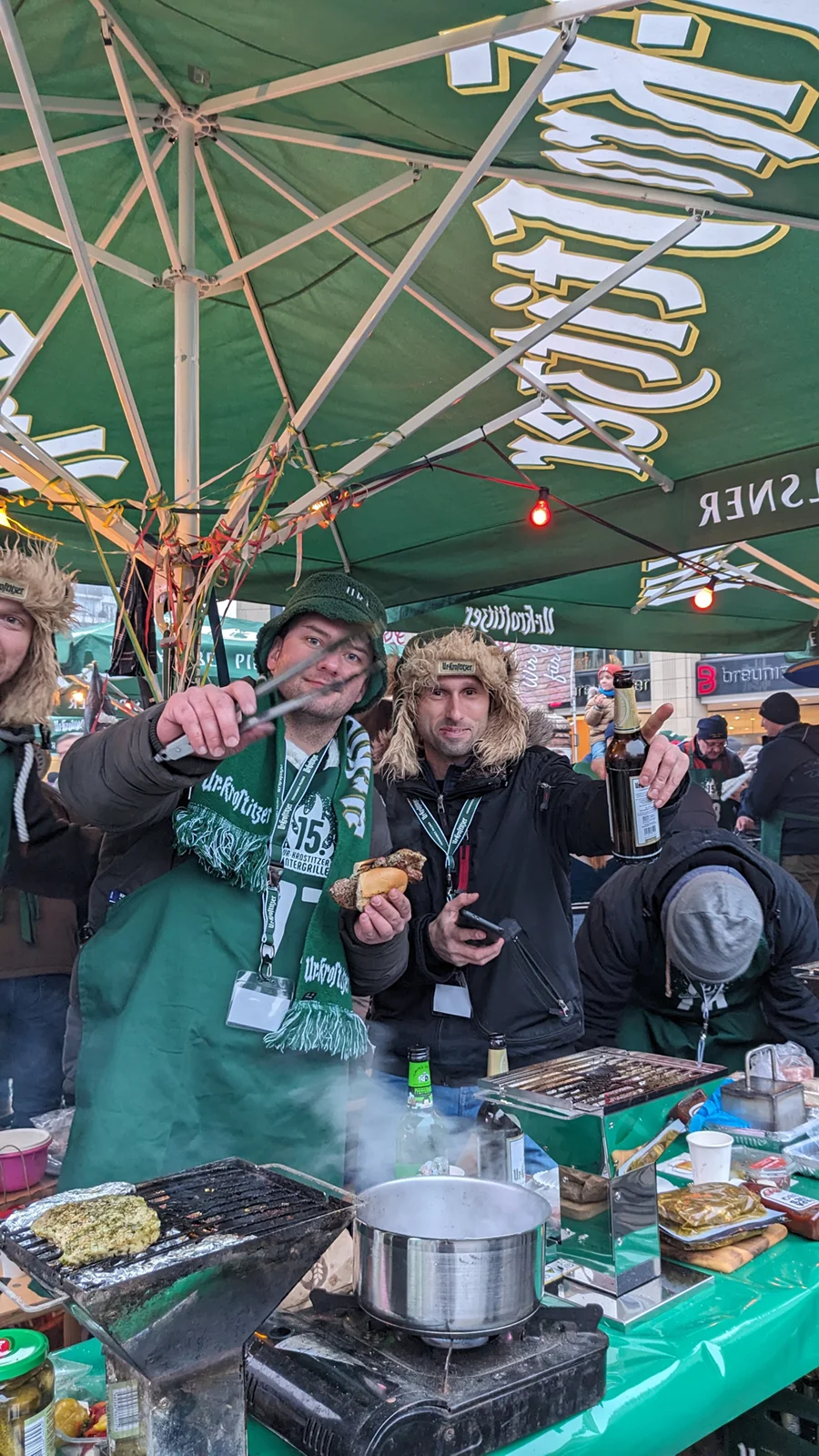 Zwei Männer in grünen Schals lachen vor einem Essensstand und halten Bier und Brötchen.