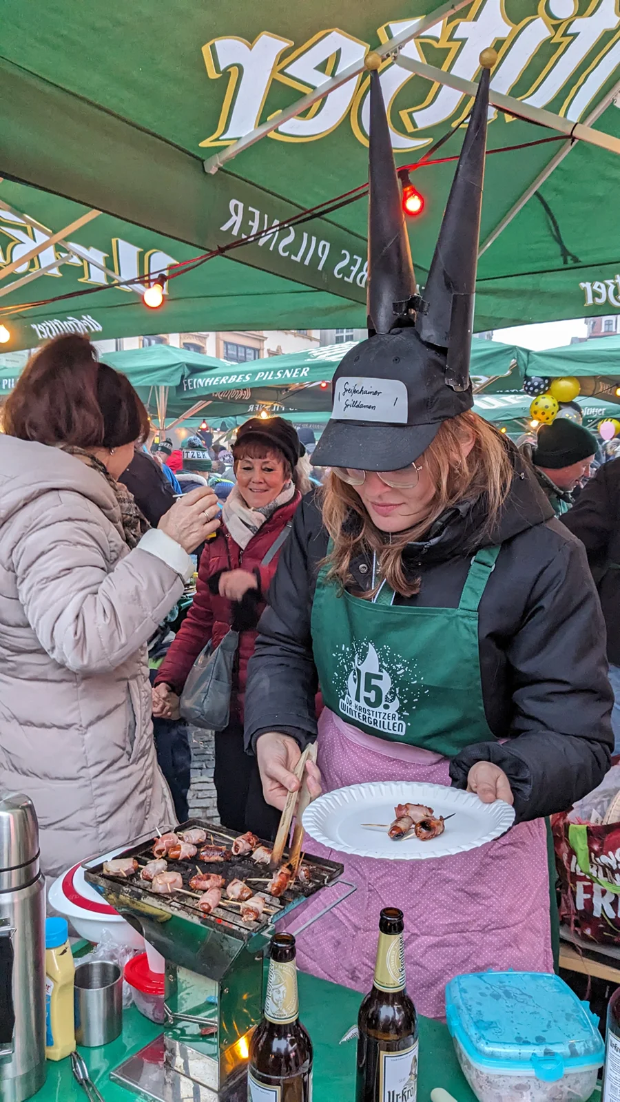 Eine Frau in einer grünen Schürze grillt Speisen auf einem kleinen Grill bei einem Outdoor-Event.