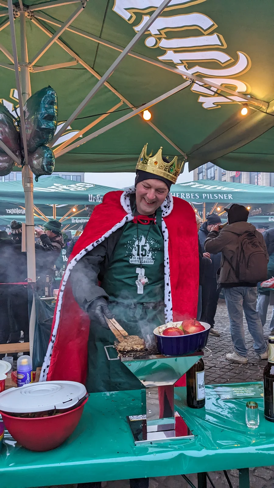 Ein Mann in Königskrone und Umhang brät Lebensmittel auf einem Marktstand.