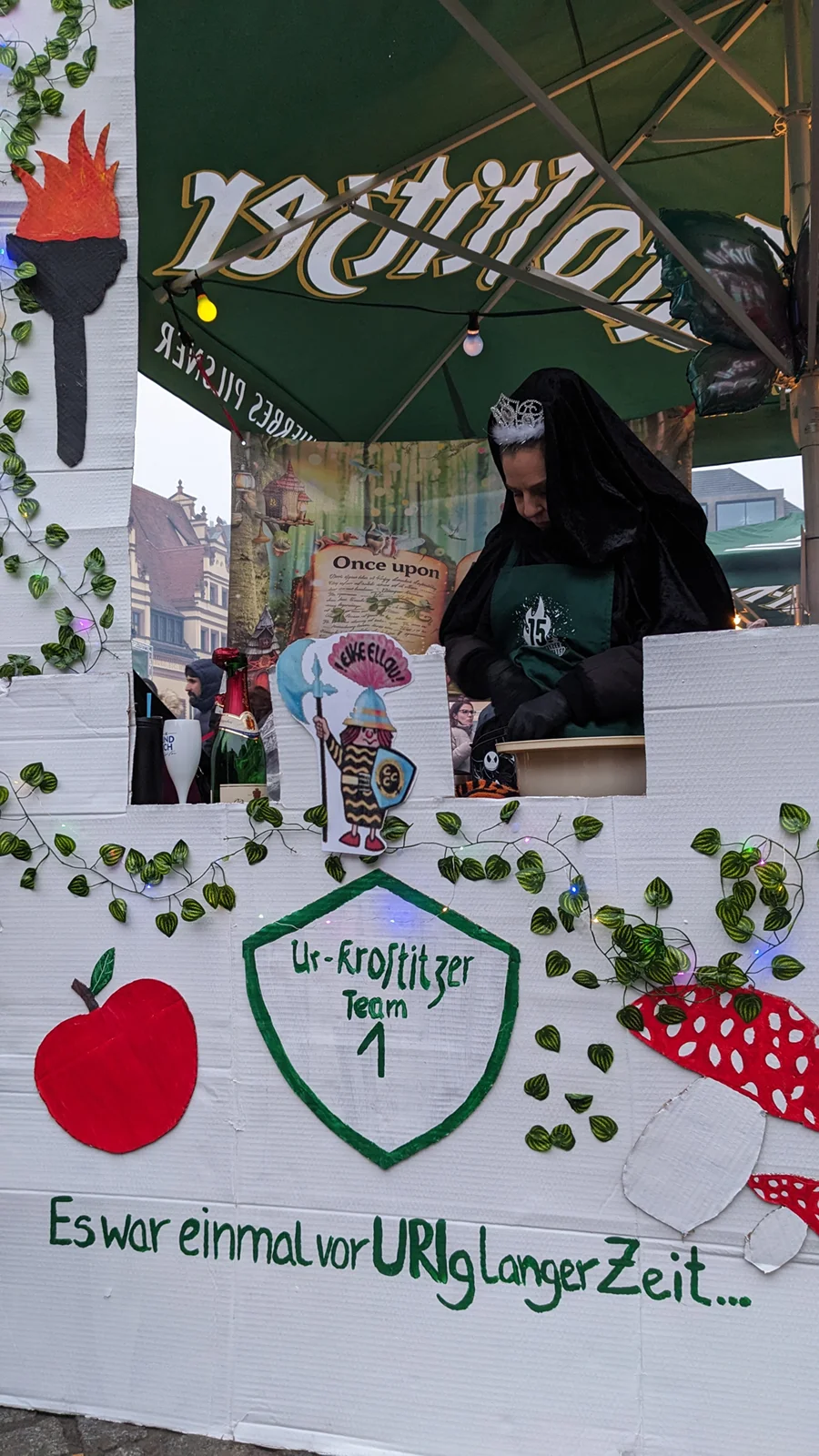 Eine Person in einem schwarzen Umhang mit Krone sitzt in einer dekorierten Marktstandnische.