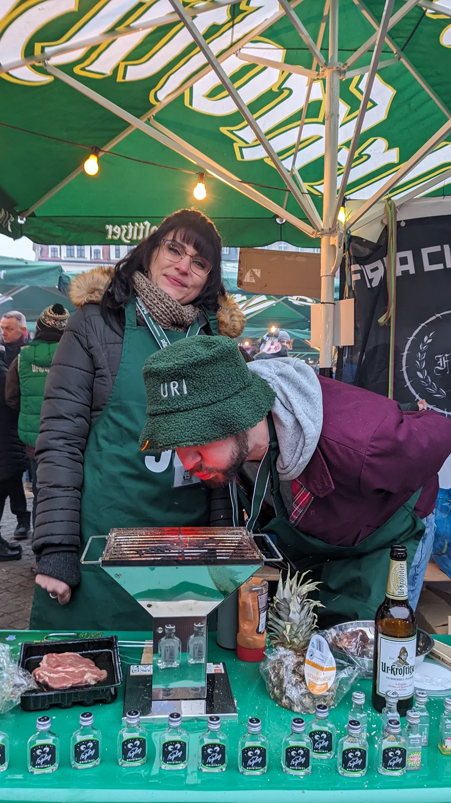Eine Frau und ein Mann stehen unter einem grünen Marktschirm an einem Stand mit Speisen und Getränken.