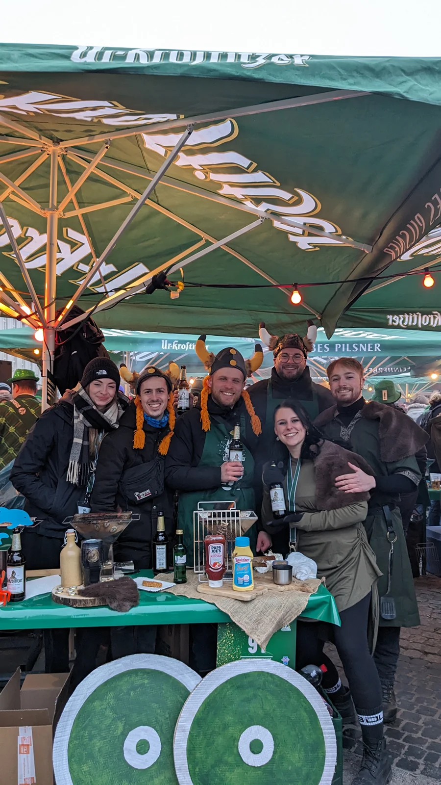 Fünf lächelnde Personen in Winterkleidung stehen an einem Marktstand. Sie tragen Hüte, die wie Hörner aussehen.