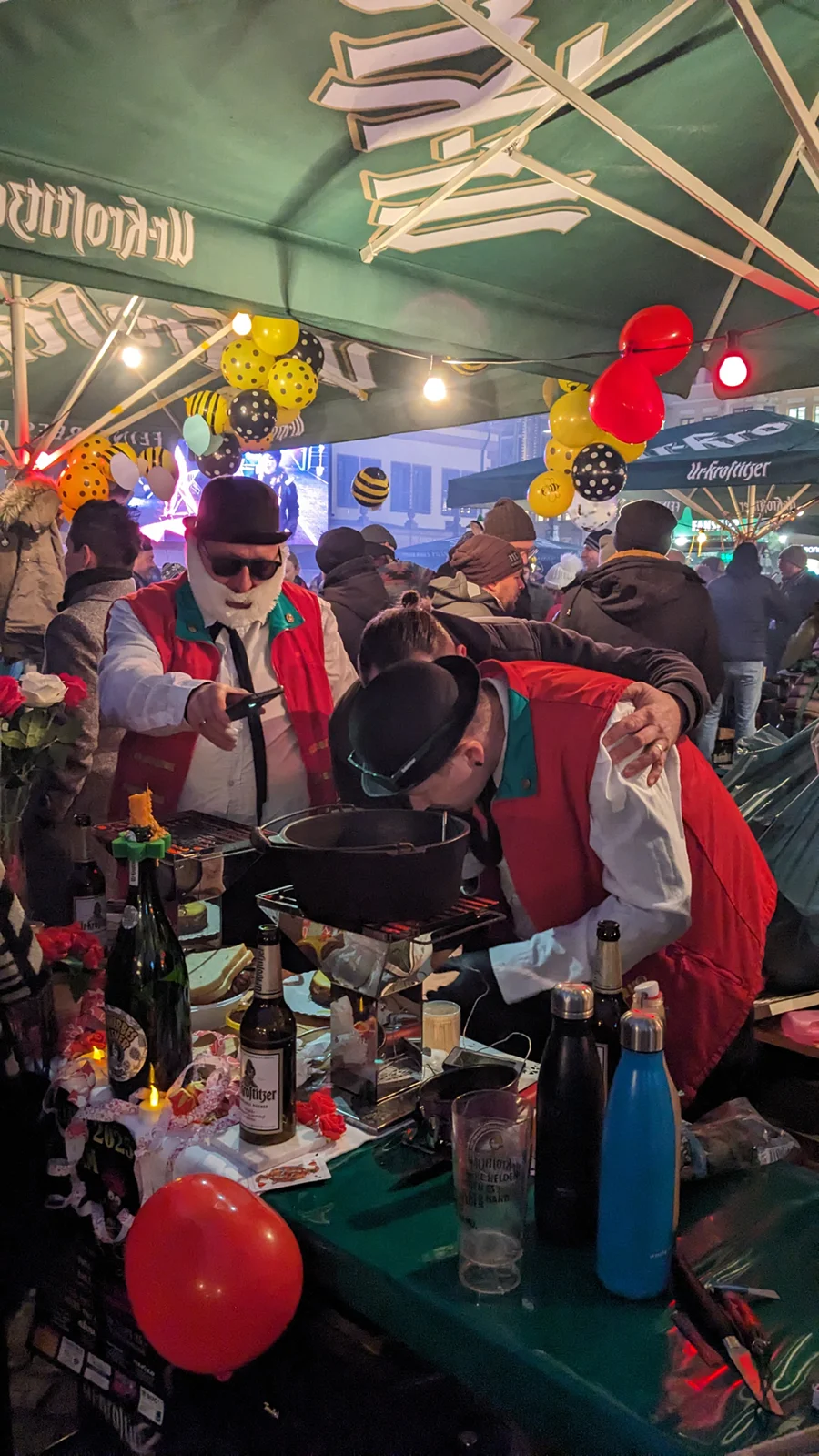 Zwei Personen bereiten Essen an einem Marktstand zu, umgeben von Ballons und Menschen.