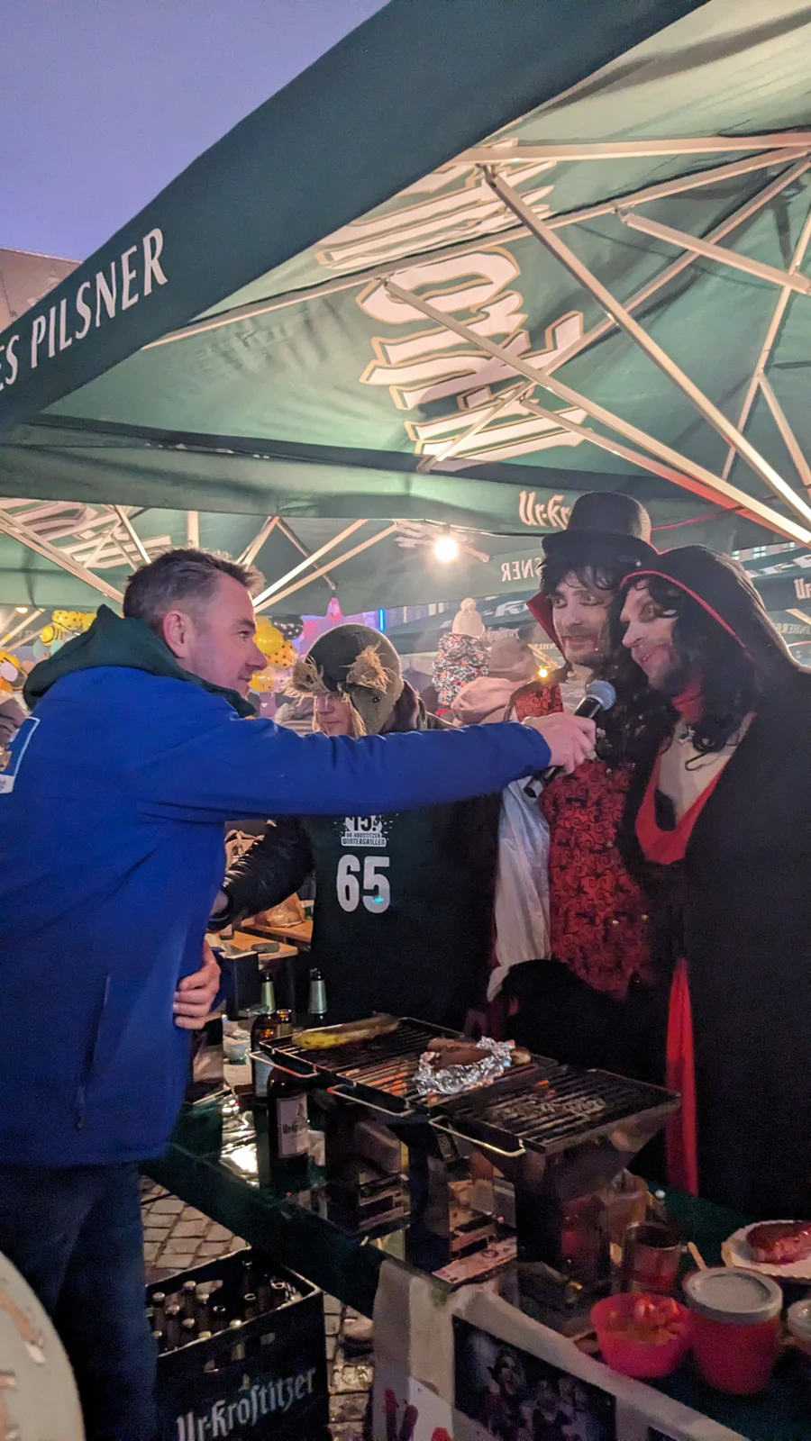 Drei Personen in Kostümen stehen an einem Grillstand bei Nacht. Eine Person hält ein Mikrofon.