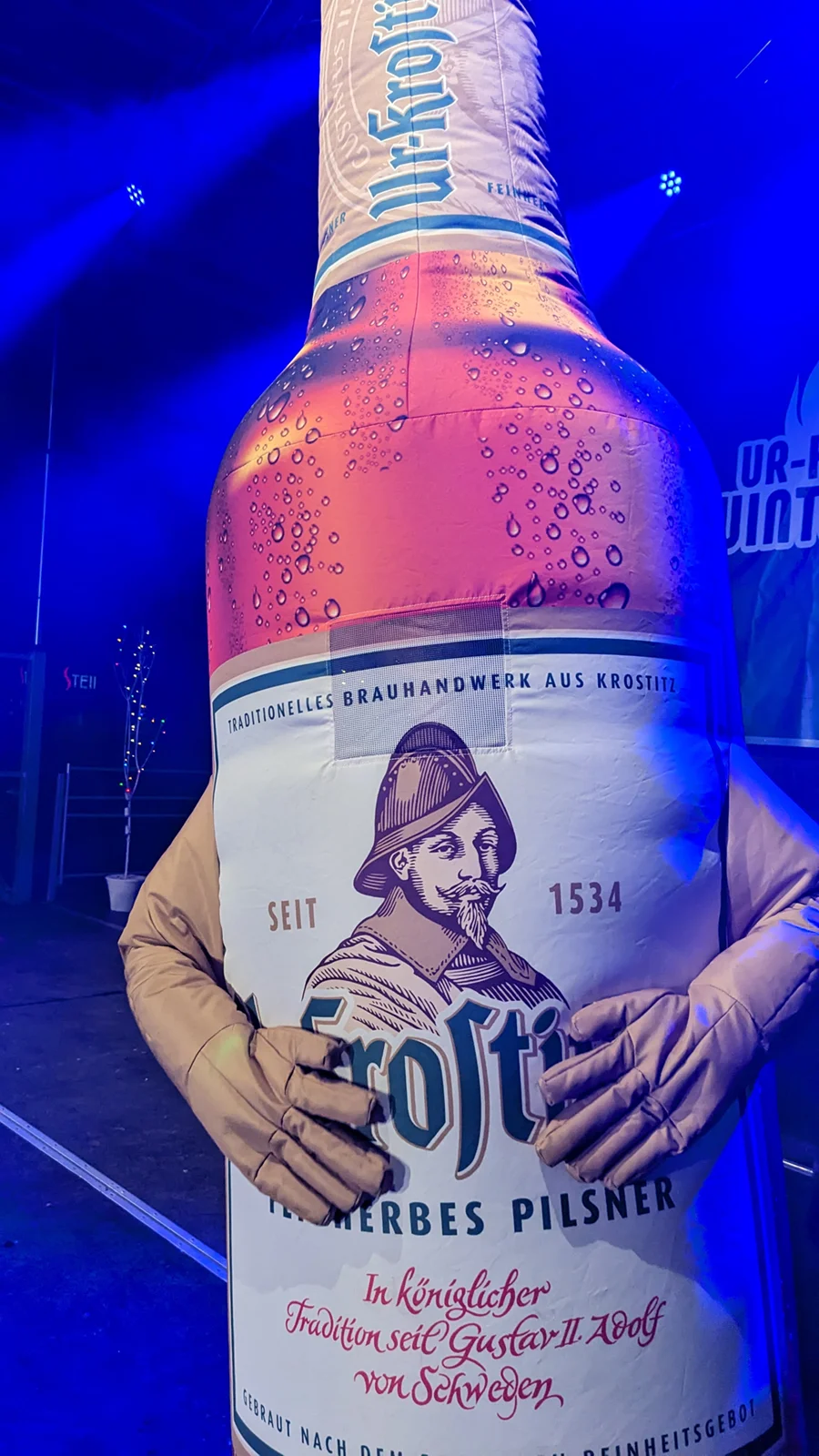 Eine Person hält ein großes aufblasbares Modell eines Bierflaschenetiketts. Das Etikett zeigt Text und ein Porträt.