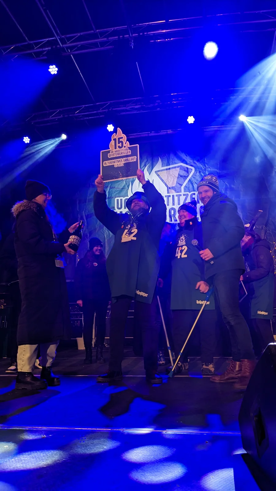 Eine Person hält stolz einen großen Preis auf einer Bühne bei einem nächtlichen Event.