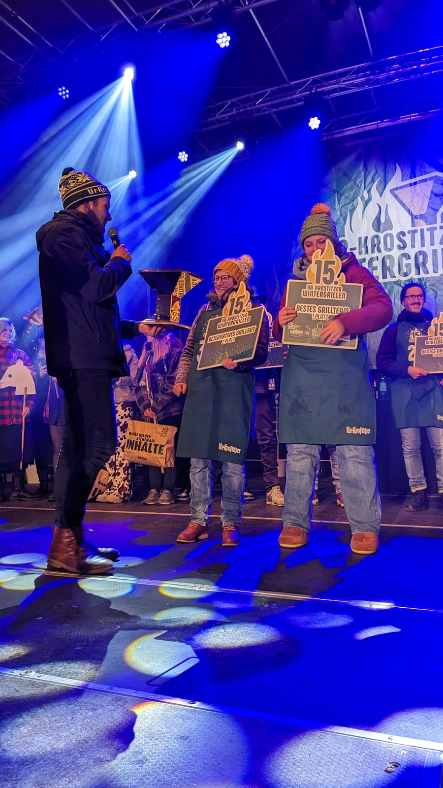 Drei Männer mit Winterkleidung halten Schilder auf einer beleuchteten Bühne.