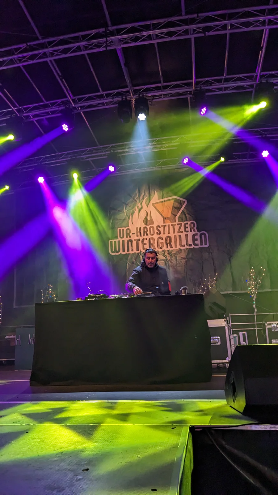 Ein DJ steht an einem Mischpult auf einer beleuchteten Bühne bei einem Event.