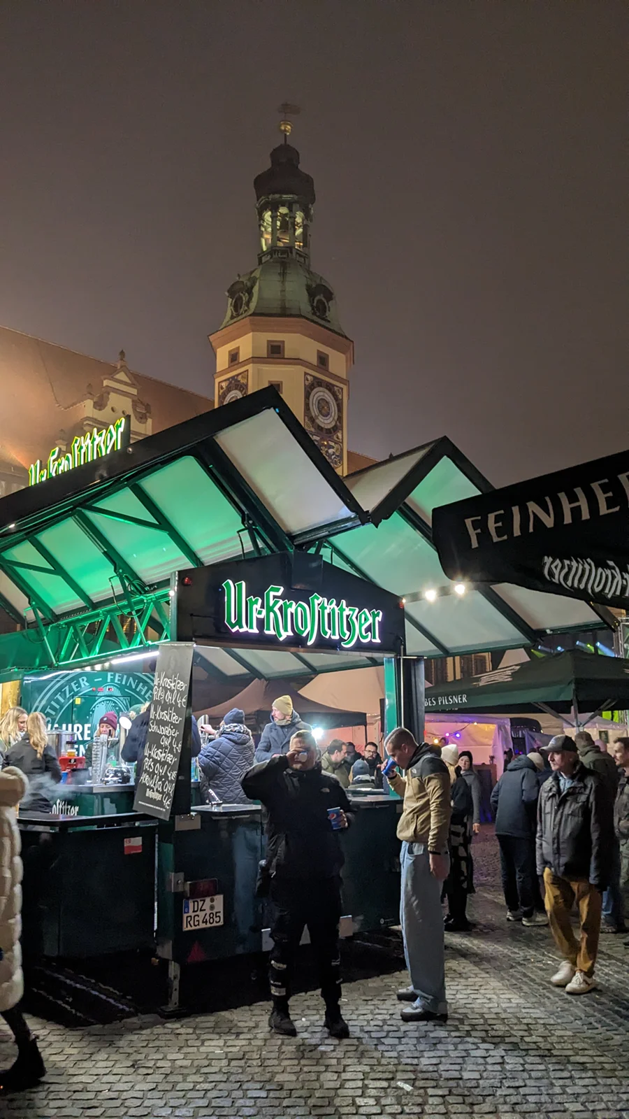 Menschen stehen in einer belebten Marktatmosphäre bei Nacht vor Getränkeständen, hinter ihnen ein historischer Turm.