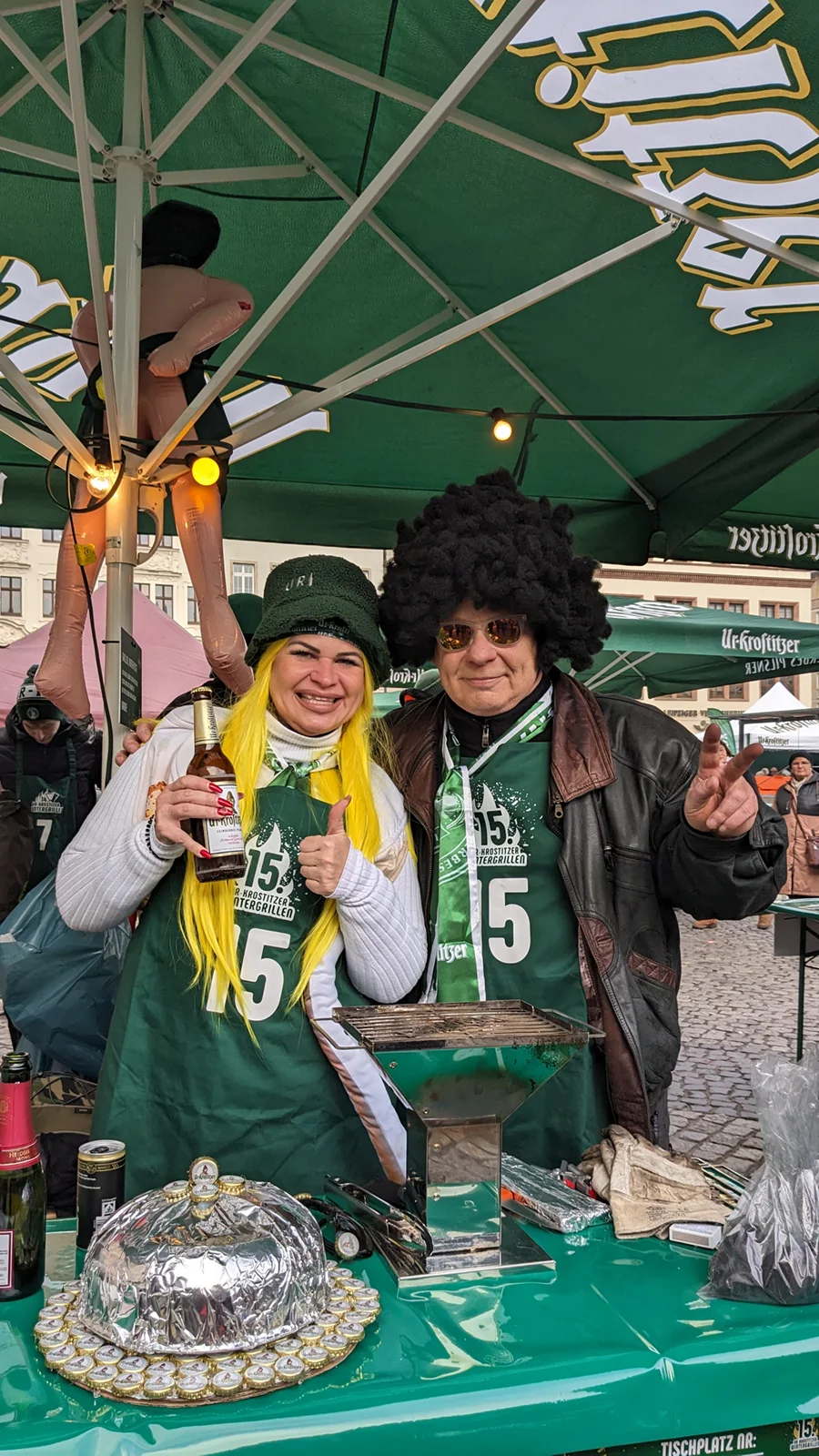 Zwei lächelnde Personen in bunten Perücken stehen unter einem grünen Schirm.