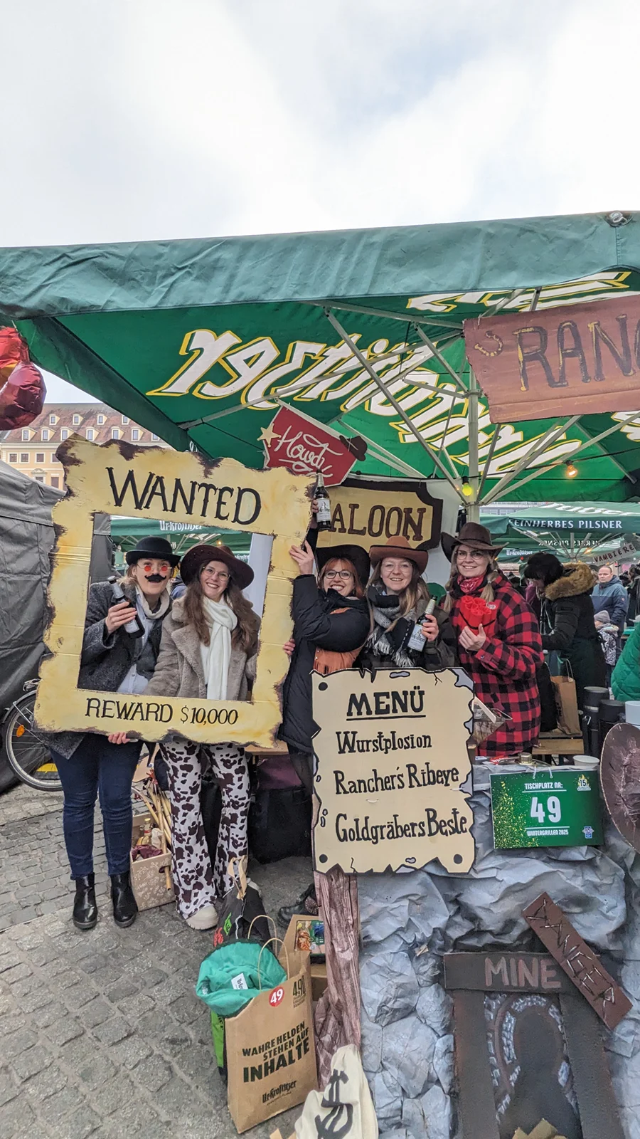 Fünf Personen posieren freudig hinter einem Rahmen mit der Aufschrift „WANTED“ auf einem Markt.