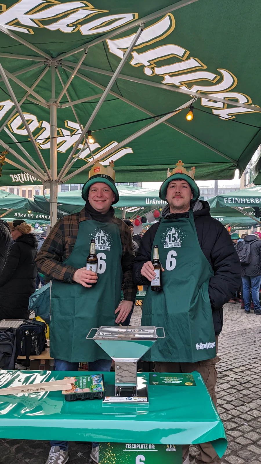 Zwei Männer in grünen Schürzen und Kronen halten Bierflaschen unter einem Marktschirm.