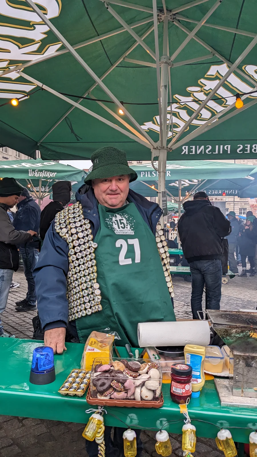 Ein Mann steht an einem Marktstand und trägt eine grüne Schürze, geschmückt mit zahlreichen Kronkorken.