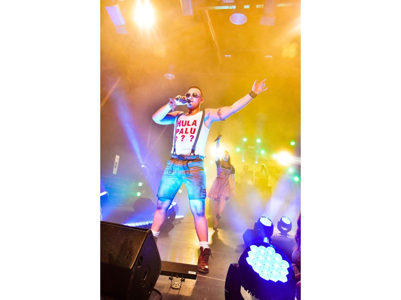 Andreas Gabalier Double Kevin steht in Lederhosen und im weißem Tanktop mit der Aufschrift Hulapalu auf einer Bühne und singt ins Mikrofon.