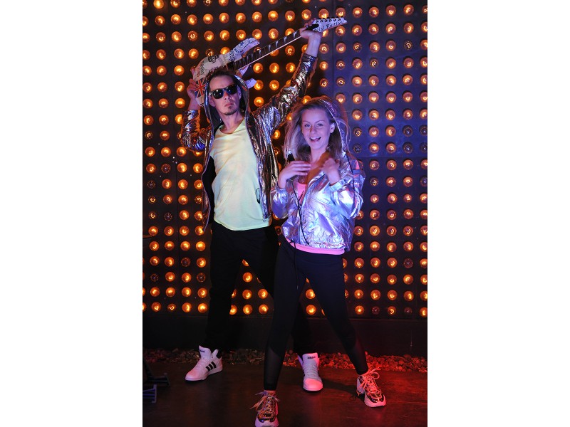  Mann und Frau stehen vor einer Wand aus leuchtenden Glühlampen in Performance Pose. Er hält eine E-Gitarre hinter und über seinem Kopf. 