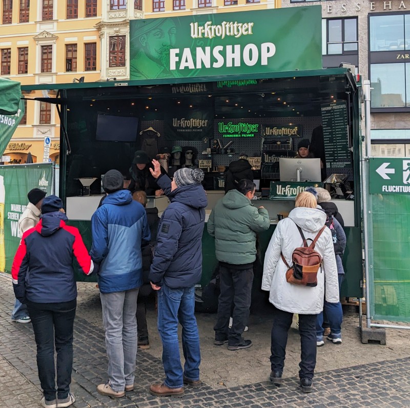 Menschen in Winterjacken stehen am Ur-Krostitzer Fanshopwagen an. Grüne Bauzaunbanner rechts und links vom Bild zu sehen.