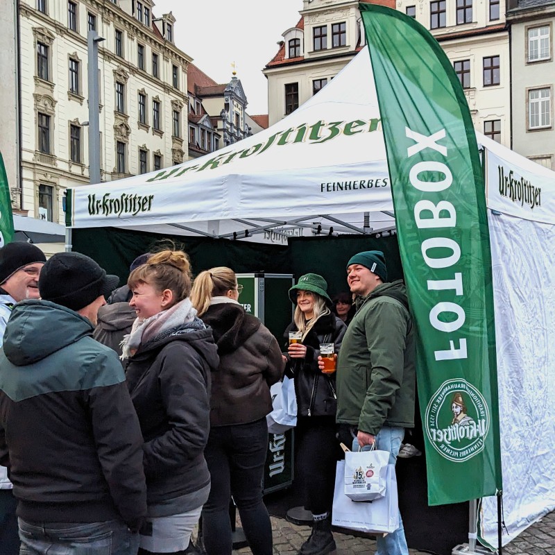 Menschen in Winterjacken stehe an einer weißen Pagode mit Ur-Krostitzer Schriftzug an. Grüne Beachflag mit Aufschrift „Fotobox“ rechts im Bild zu sehen.