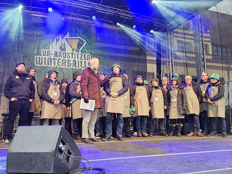 Mehre Menschen in Grillschürzen stehen auf einer Bühne und werden von einem Moderator befragt. Banner mit dem Logo „15. Ur-Krostitzer Wintergrillen“ im Hintergrund.