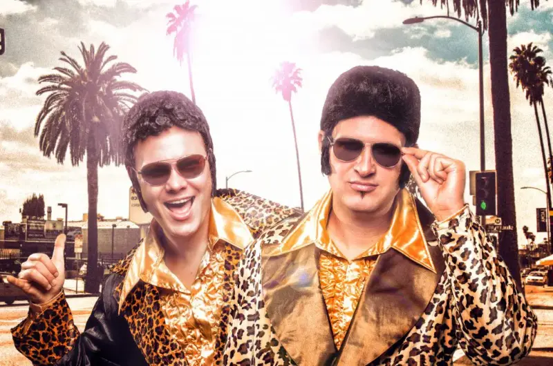 Zwei Männer in Leoparden-Print-Jacketts und 70er Jahre Frisuren lachen in die Kamera. Im Hintergrund Palmen.