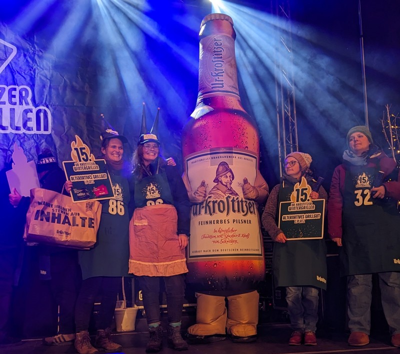 Vier Personen in grünen Grillschürzen stehen auf einer Bühne. In der Mitte eine Ur-Krostitzer Flaschen Maskottchen.