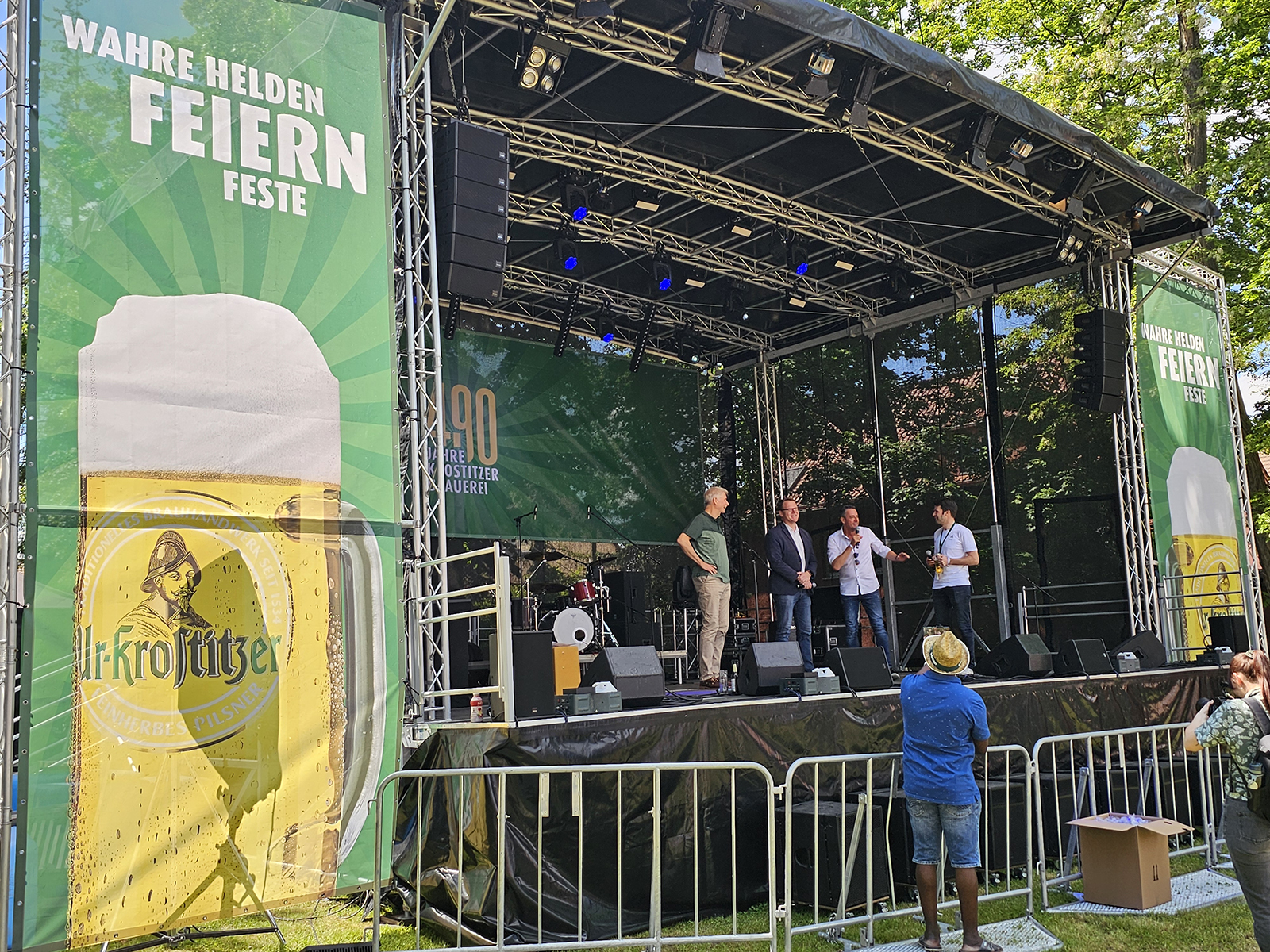 Vier Männer stehen auf einer Bühne und sprechen, beobachtet von einem Publikum im Freien.