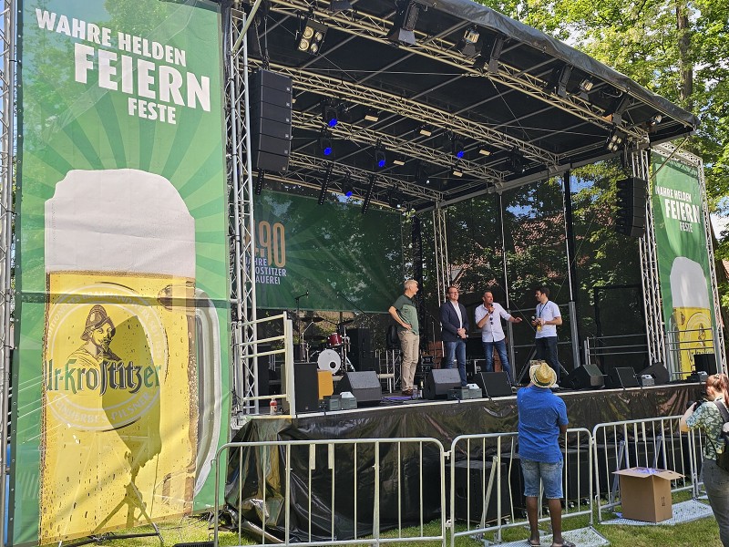 Vier Männer stehen auf einer Bühne und sprechen, beobachtet von einem Publikum im Freien.