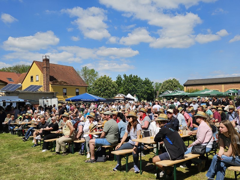 Eine große Menschenmenge sitzt bei sonnigem Wetter an Bierzeltgarnituren. Viele tragen Strohhüte.