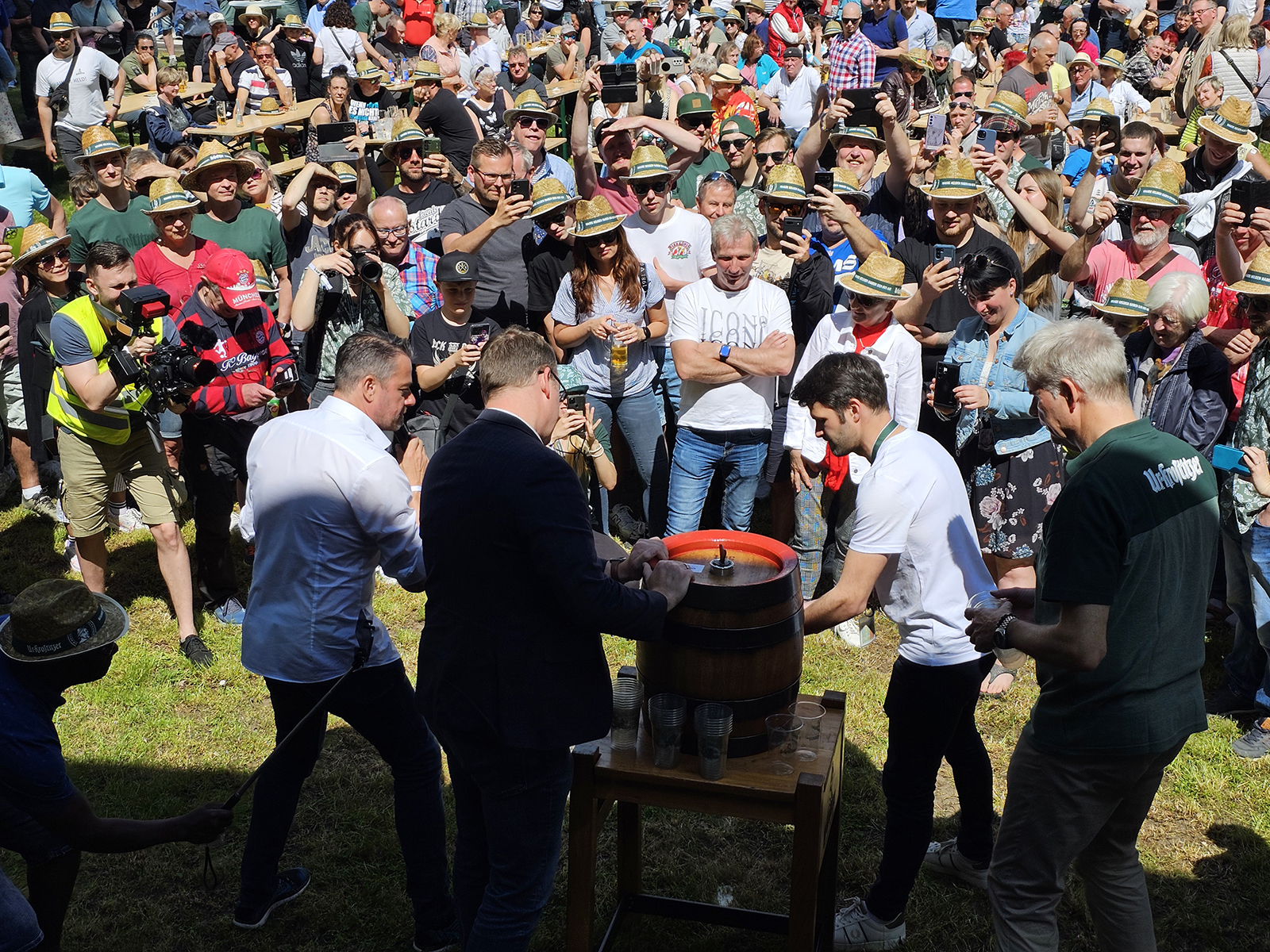Eine Gruppe Menschen mit Strohhüten steht im Freien und umringt eine Bierfasszapfung.