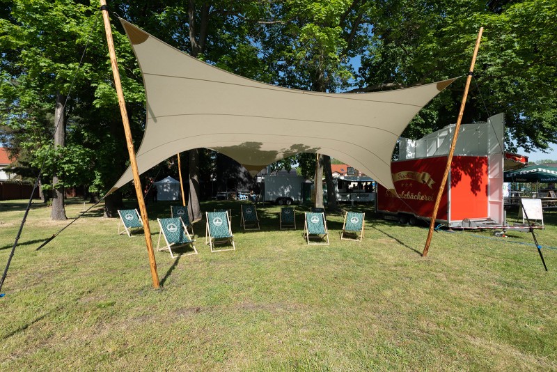 Fünf Liegestühle stehen unter einem großen, beige gespannten Sonnensegel auf einer Wiese.