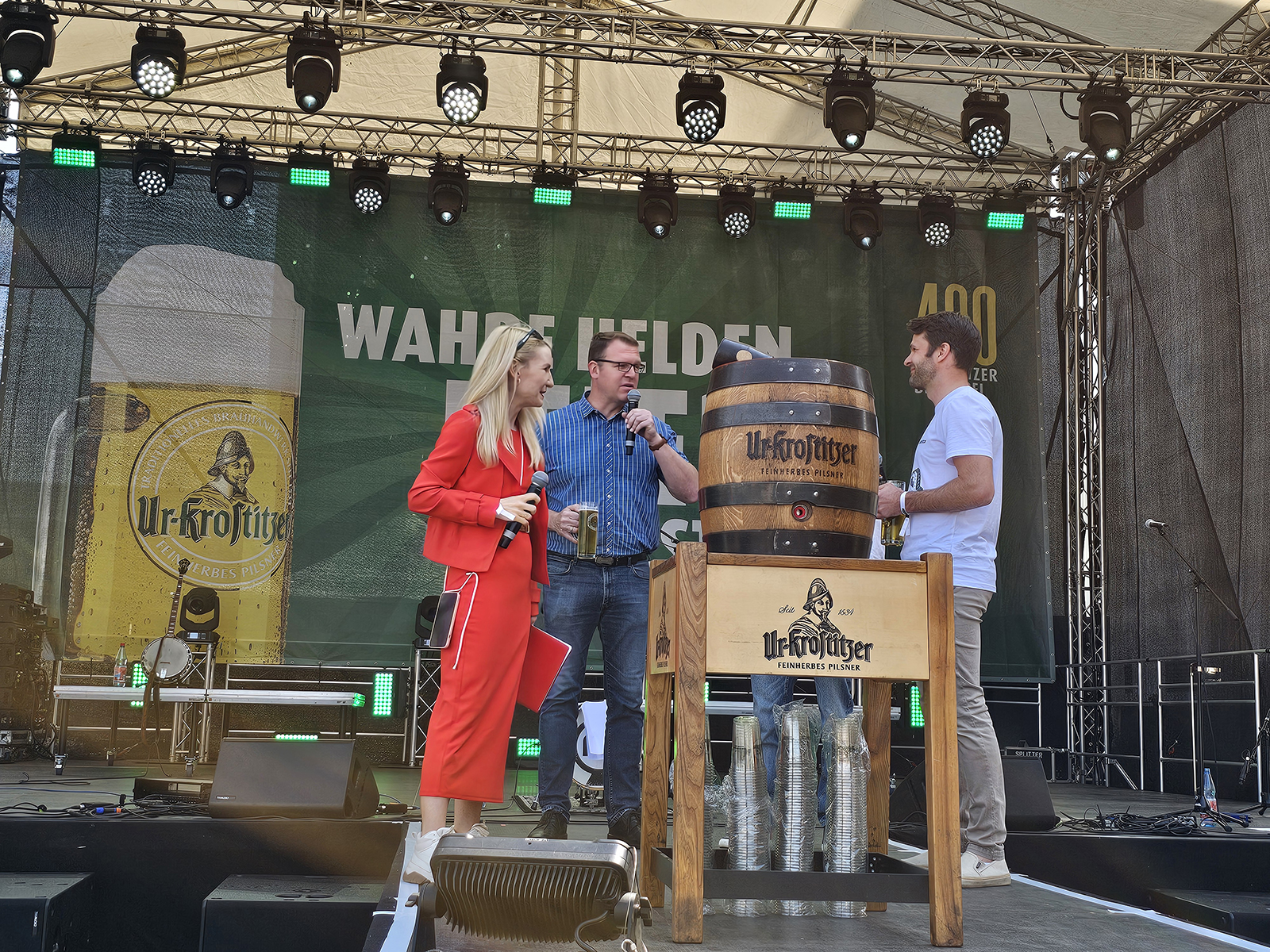 Drei Personen stehen auf einer Bühne neben einem großen Bierfass und sprechen in Mikrofone.