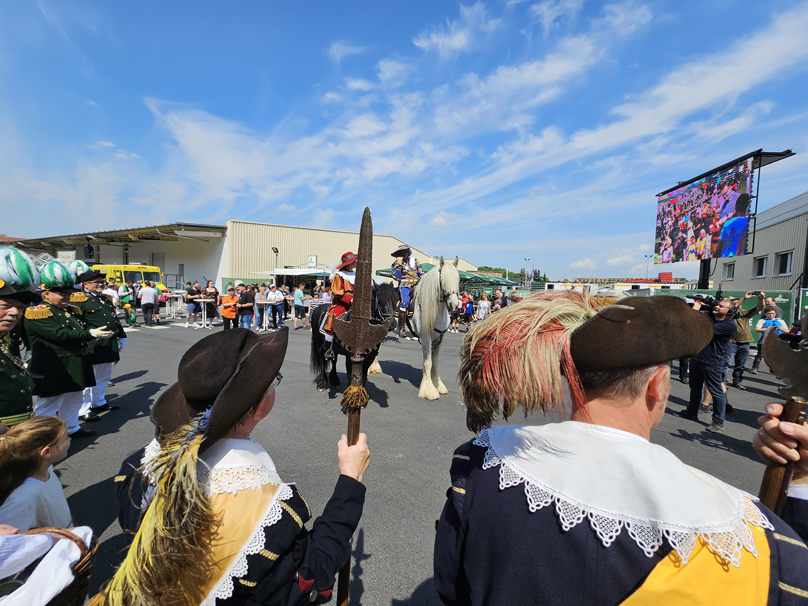Menschen in traditioneller Kleidung beobachten eine Parade mit einem Pferd unter blauem Himmel.