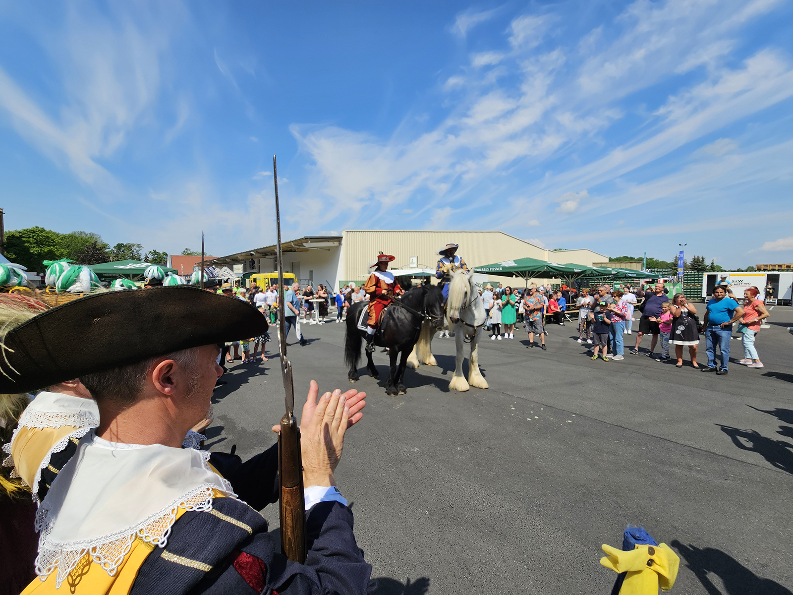 Ein Mann in historischer Kleidung schaut zwei Reitern auf Pferden zu. Der Himmel ist blau mit Wolken.