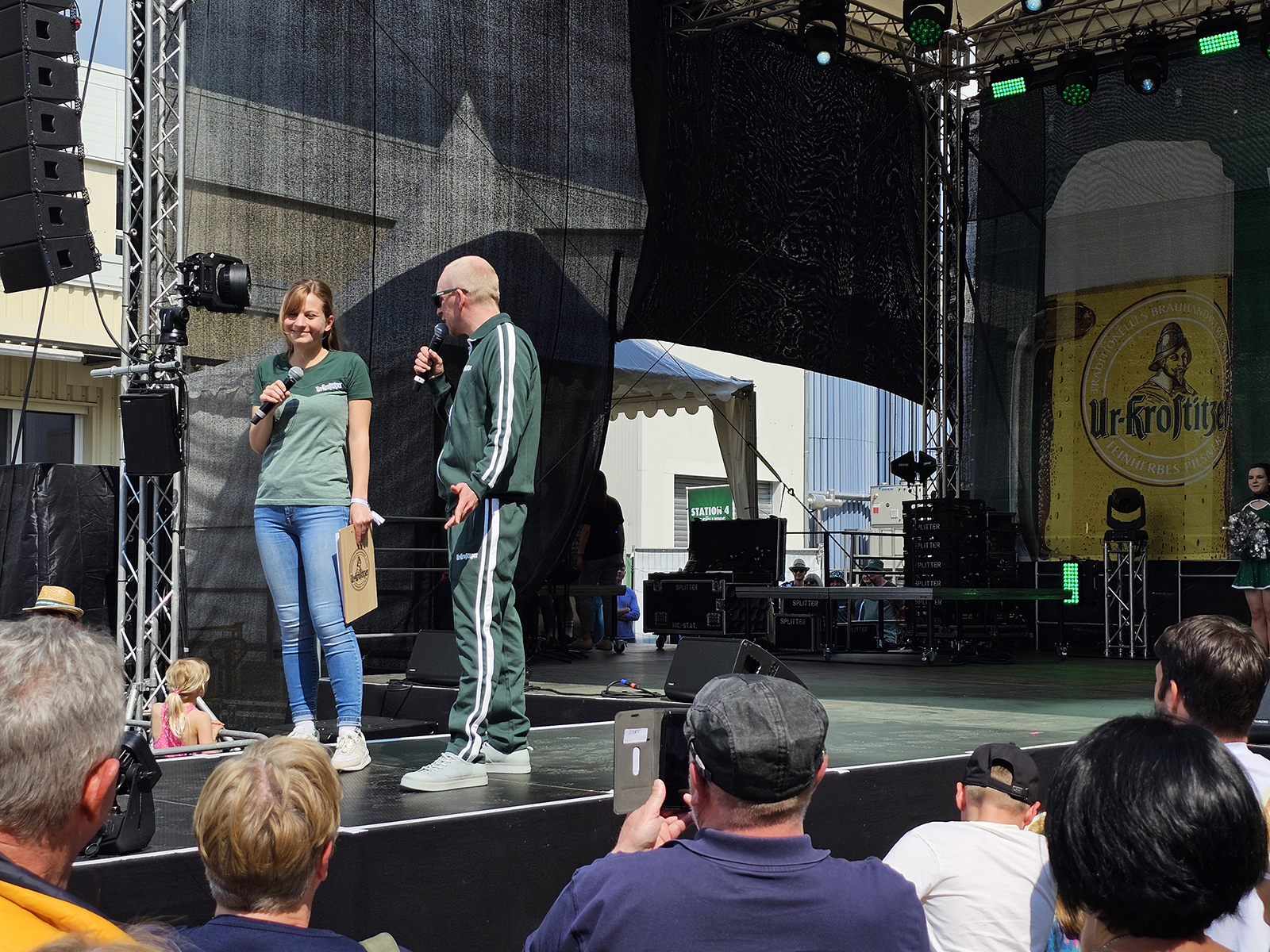 Zwei Personen halten Mikrofone und stehen auf einer Bühne bei einem Open-Air-Event.