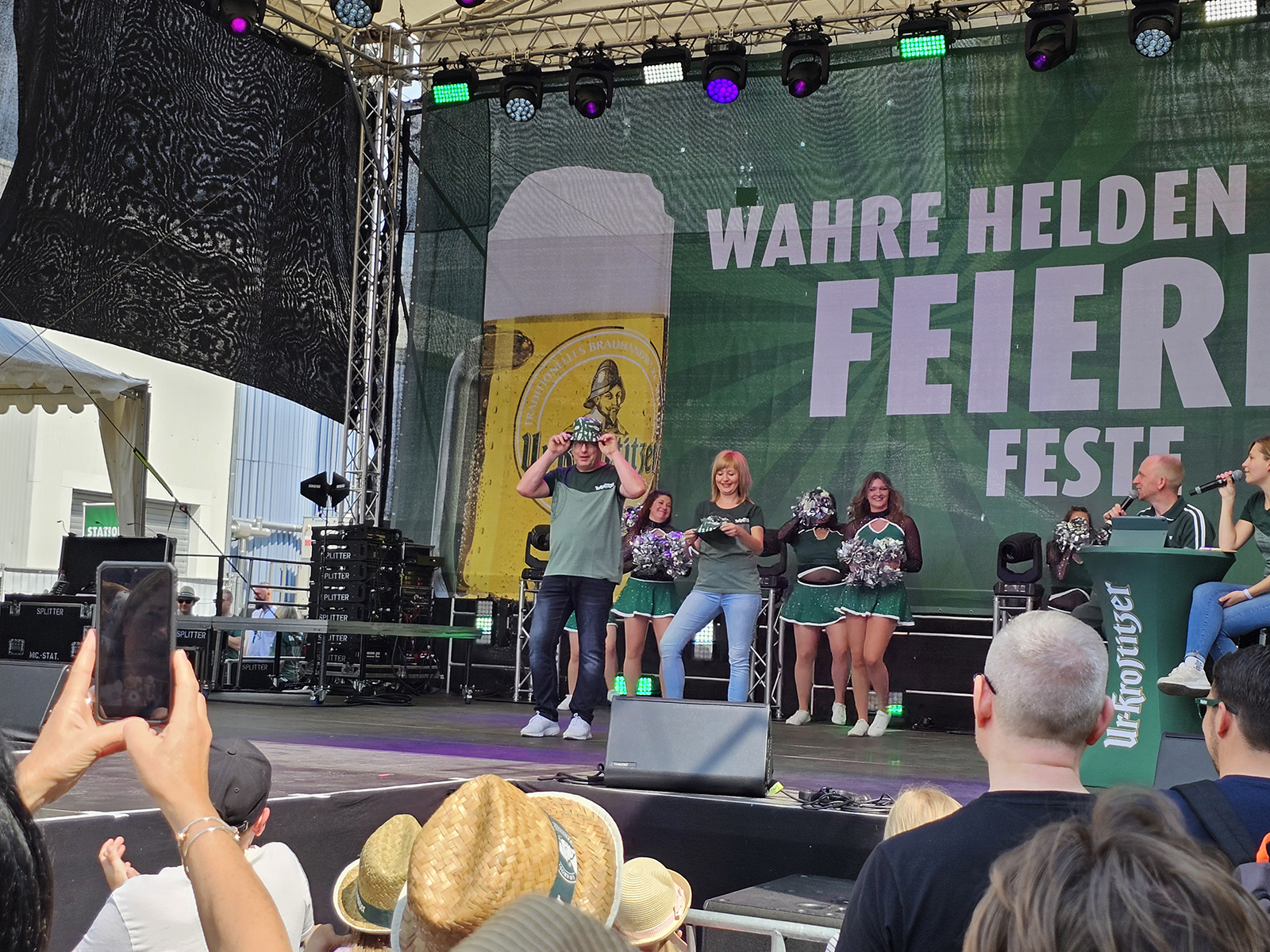 Mehrere Personen stehen auf einer Bühne bei einem öffentlichen Event. Einige tanzen, während andere zuschauen.