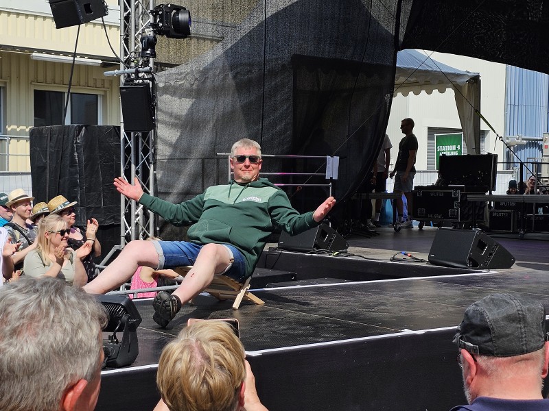 Ein Mann sitzt lässig auf einer Bühne und gestikuliert, umgeben von Publikum und Bühnentechnik.