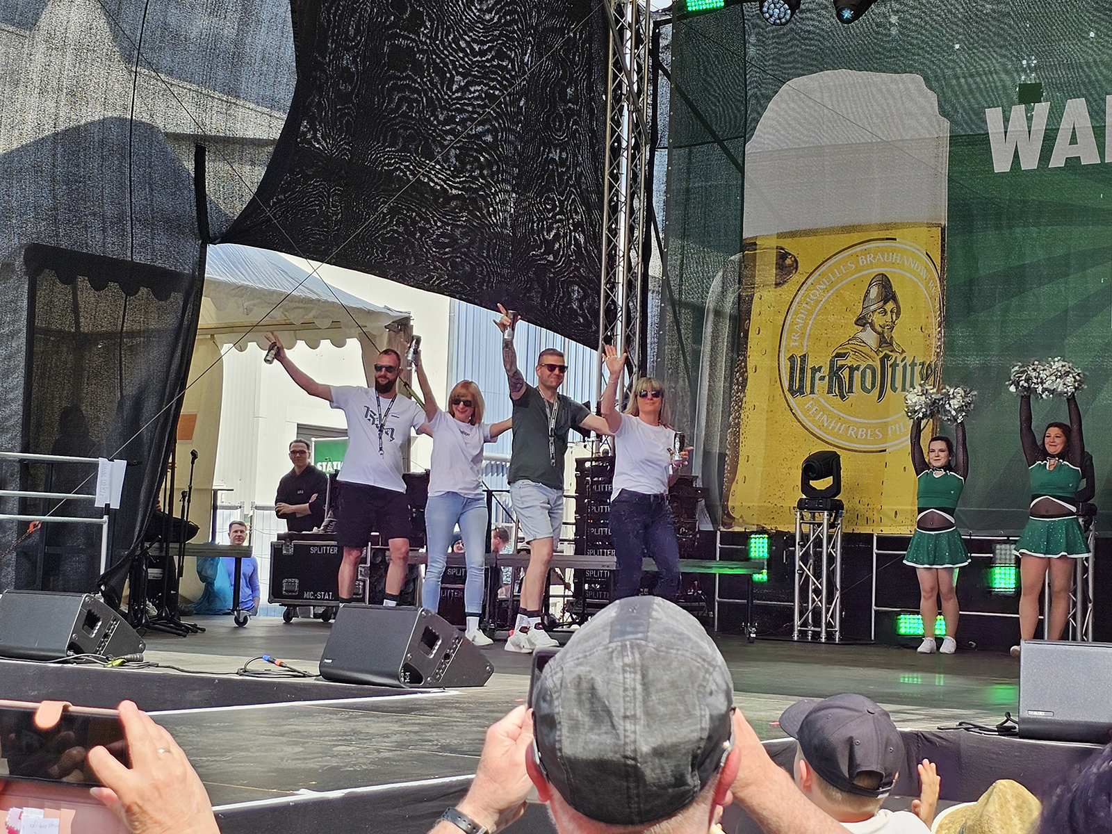 Fünf Erwachsene feiern auf einer Bühne, flankiert von vier Cheerleadern.