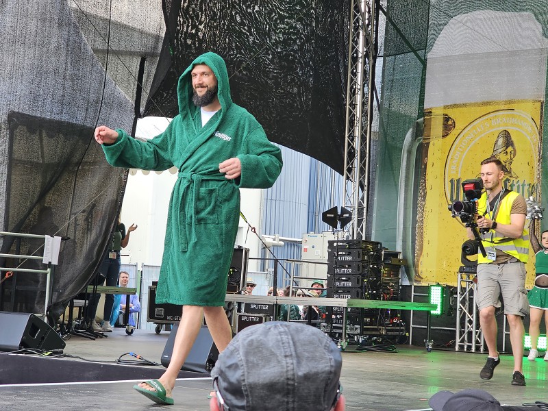 Ein Mann in einem grünen Bademantel steht lächelnd auf einer Bühne.