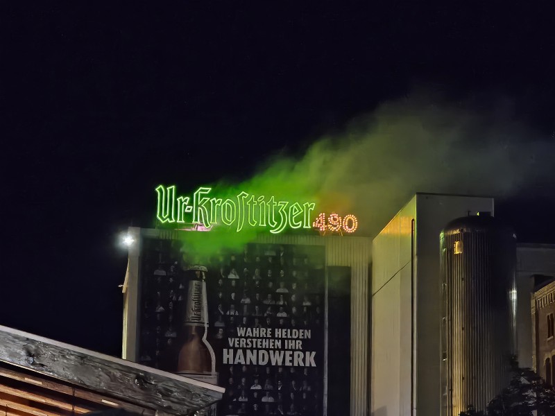 Neonleuchtschrift auf einem Gebäudedach wird von grünem Nebel umhüllt. Ein Wandplakat zeigt eine Bierflasche.