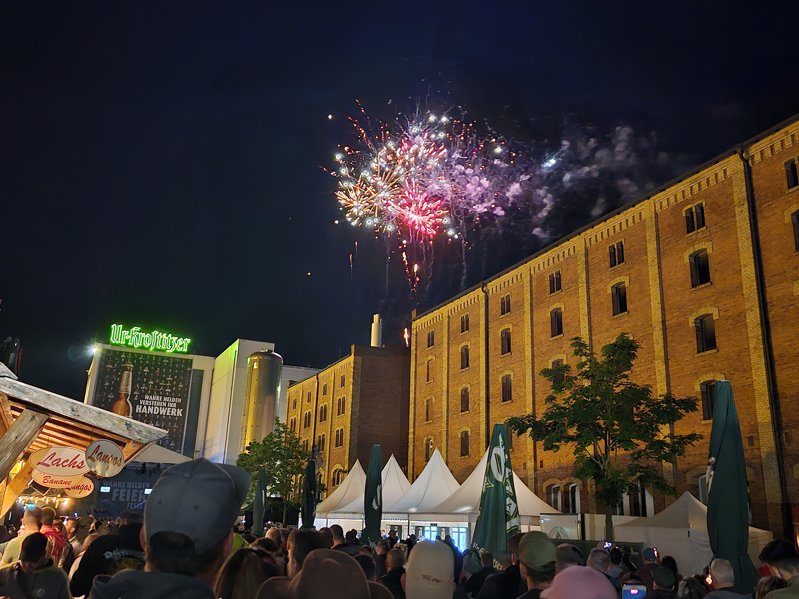 Feuerwerk erleuchtet den Nachthimmel über einem belebten Festival neben einem großen Gebäude.