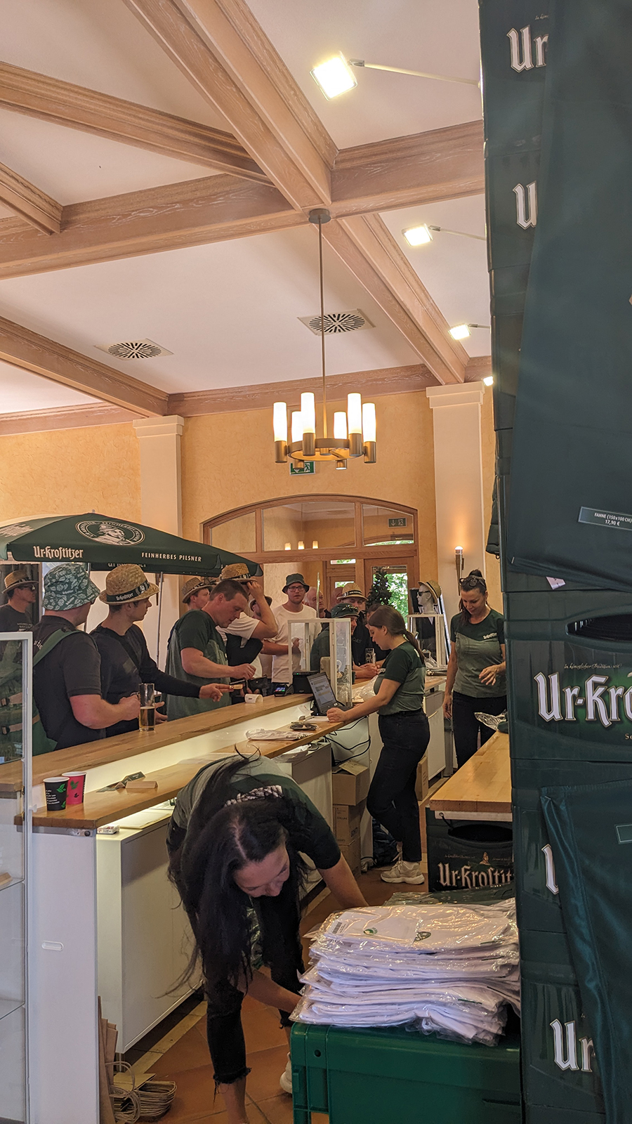 Mehrere Personen stehen an einer Theke in einem Raum mit Holzdecke, einige tragen grüne Hüte. Eine Frau packt T-Shirts vor einem Werbestand aus.