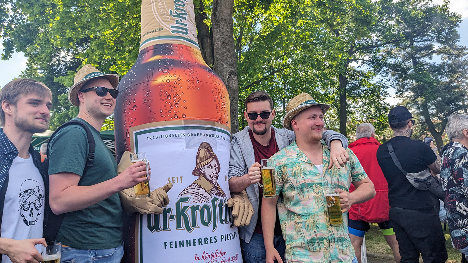 Vier Männer stehen lächelnd vor einer großen Bierflaschenattrappe im Park.