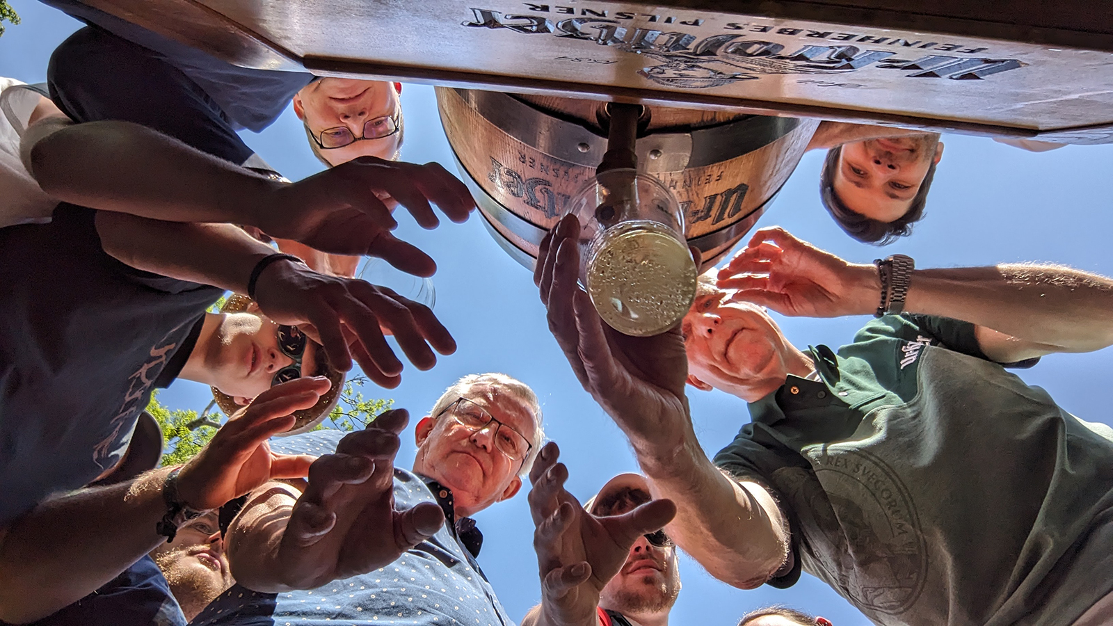 Mehrere Personen greifen nach einem Glas Bier, das von unten fotografiert wird.