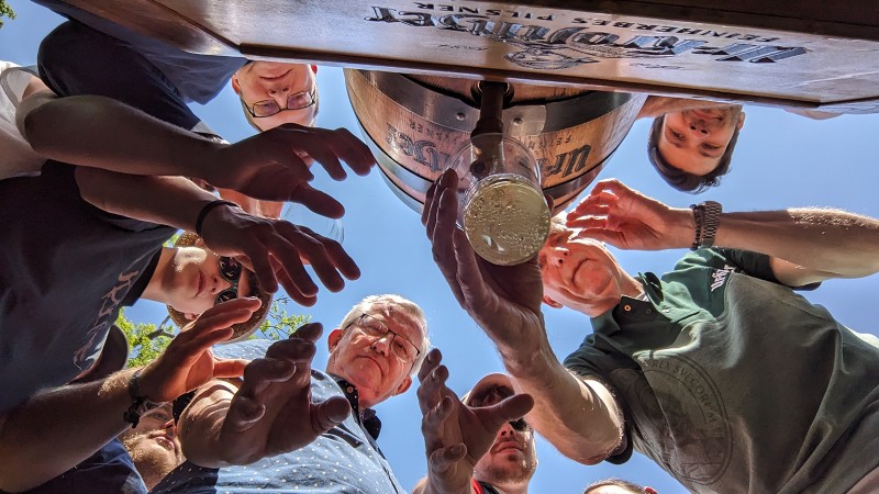 Mehrere Personen greifen nach einem Glas Bier, das von unten fotografiert wird.