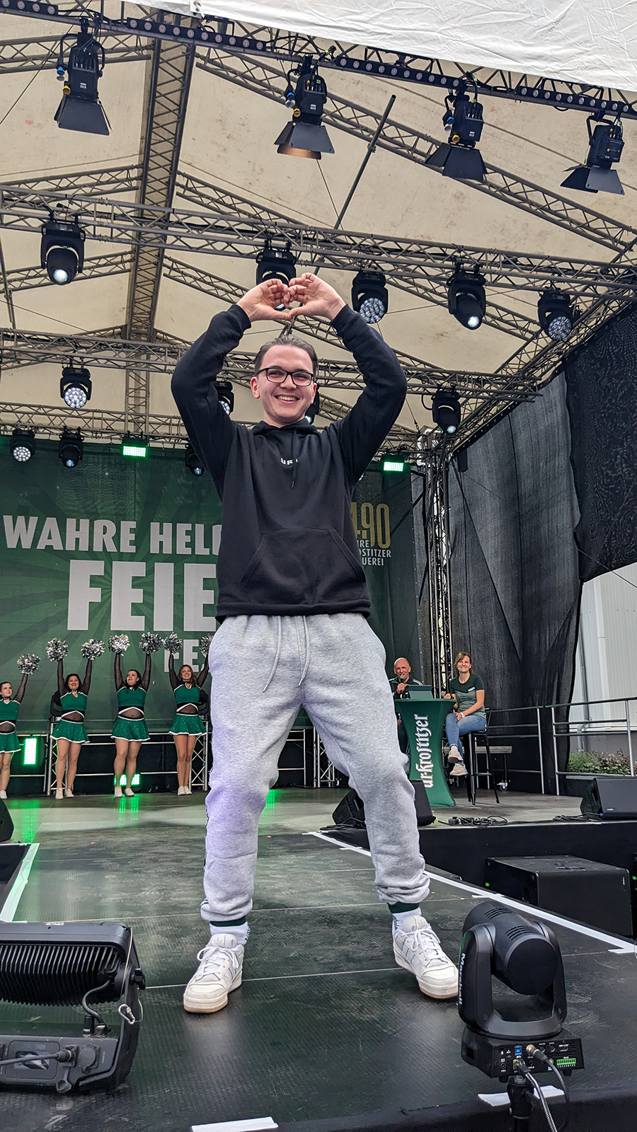 Ein junger Mann steht auf einer Bühne und formt ein Herz mit den Händen über seinem Kopf. Hinter ihm sind Cheerleader und Bühnenbeleuchtung zu sehen.