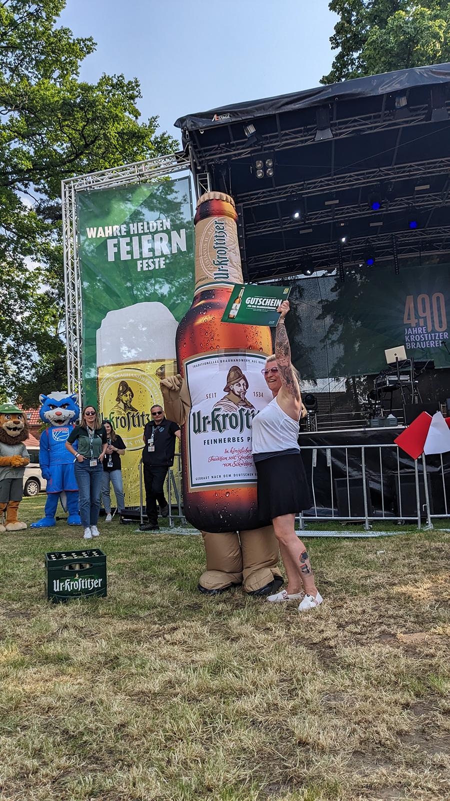 Eine Frau umarmt eine große aufblasbare Flasche auf einer Veranstaltungsbühne, im Hintergrund ist Gras.