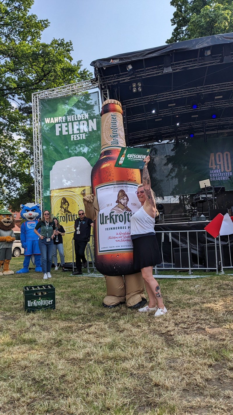Eine Frau umarmt eine große aufblasbare Flasche auf einer Veranstaltungsbühne, im Hintergrund ist Gras.